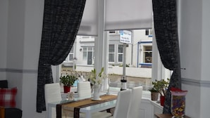 A room with a large window, white curtains, a dining table with chairs, and potted plants. - The Cameo (Blackpool)