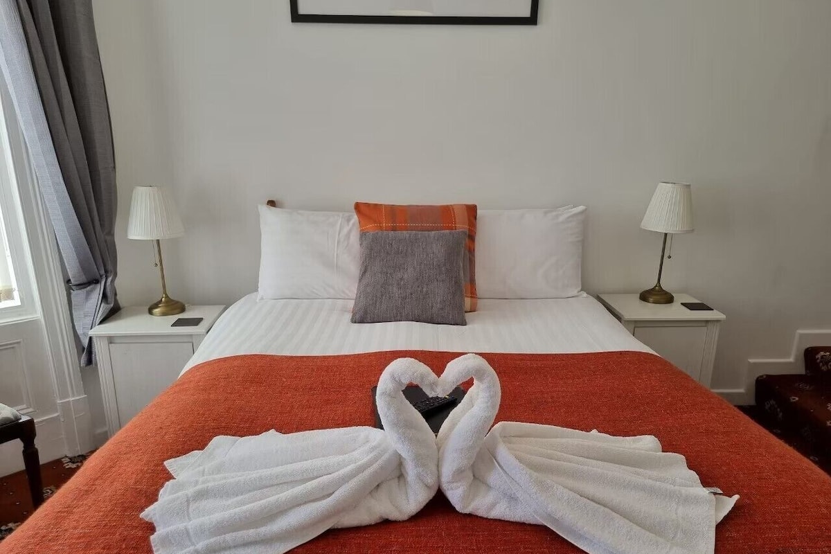 A hotel room with a bed, two bedside tables, a nightstand, a red throw blanket, and towels arranged in a heart shape.