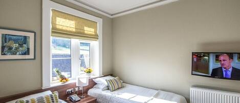 A hotel room with two beds, a window with blinds, a television on the wall, and a radiator.