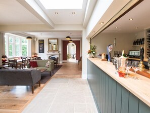 Uma sala de estar ampla com lareira, um espaço bar com copos e garrafas de vinho, e vista para o exterior através de grandes janelas.