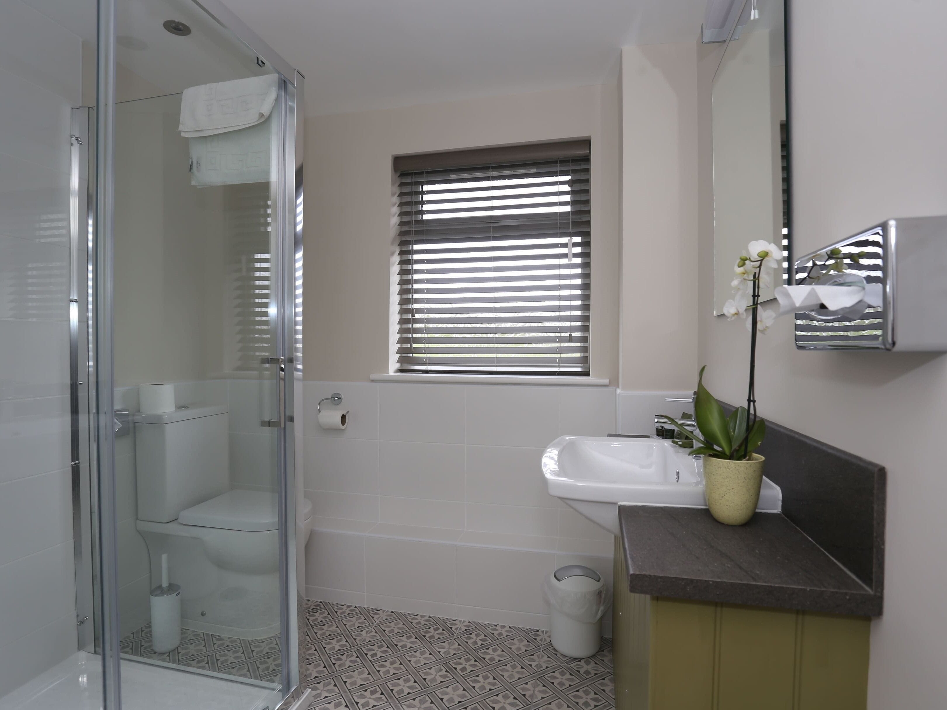 A bathroom with a glass shower enclosure, a toilet, a sink, and a window with blinds.