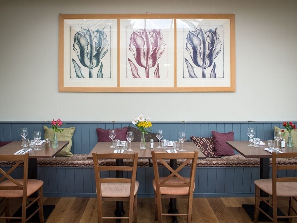 Ein Essbereich mit Holztischen und -stühlen, einer blauen Bank und gerahmten Blumenbildern an der Wand.