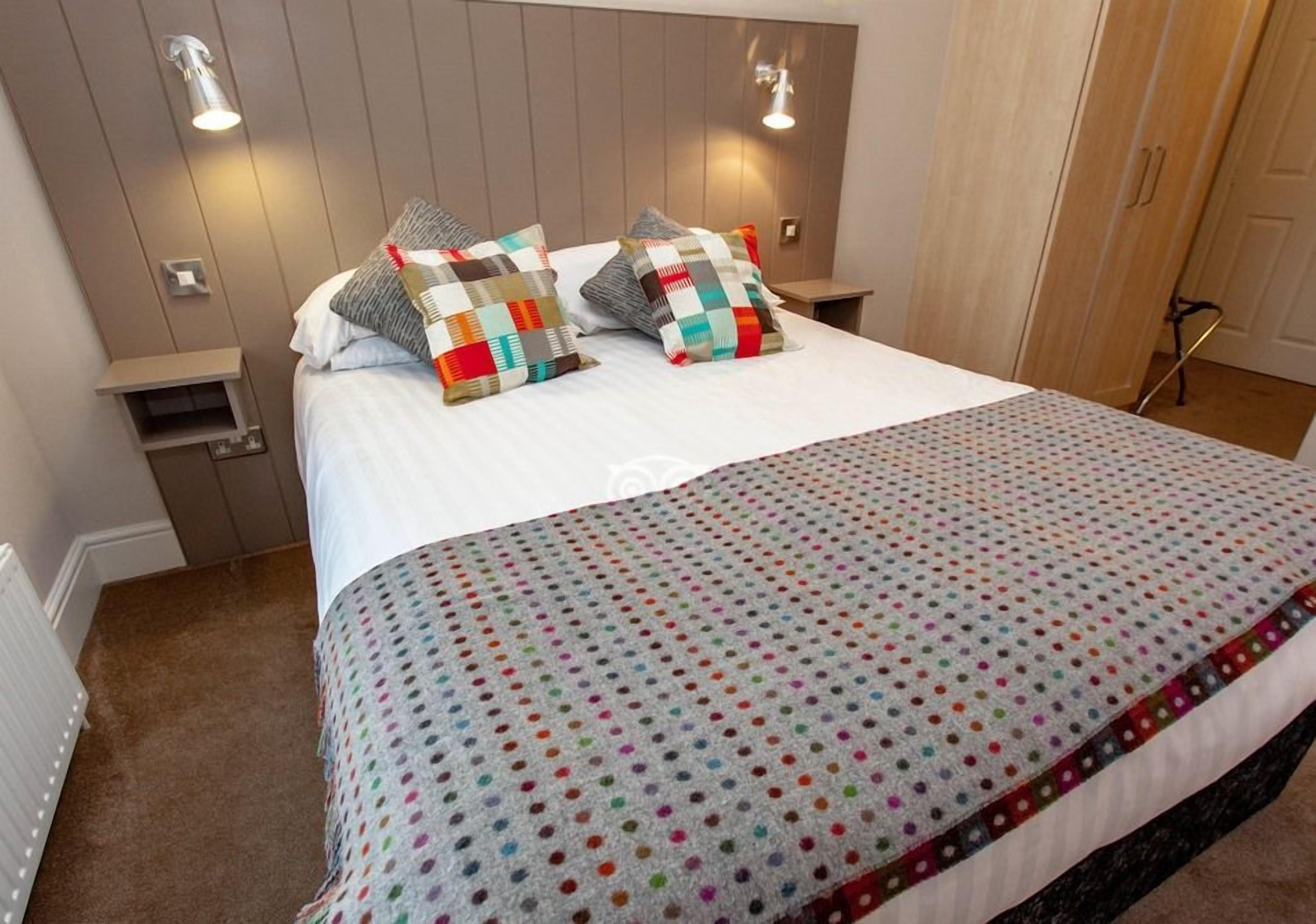 A neatly made bed with a colorful polka dot blanket and two decorative pillows, a bedside table, and a wall-mounted light fixture.