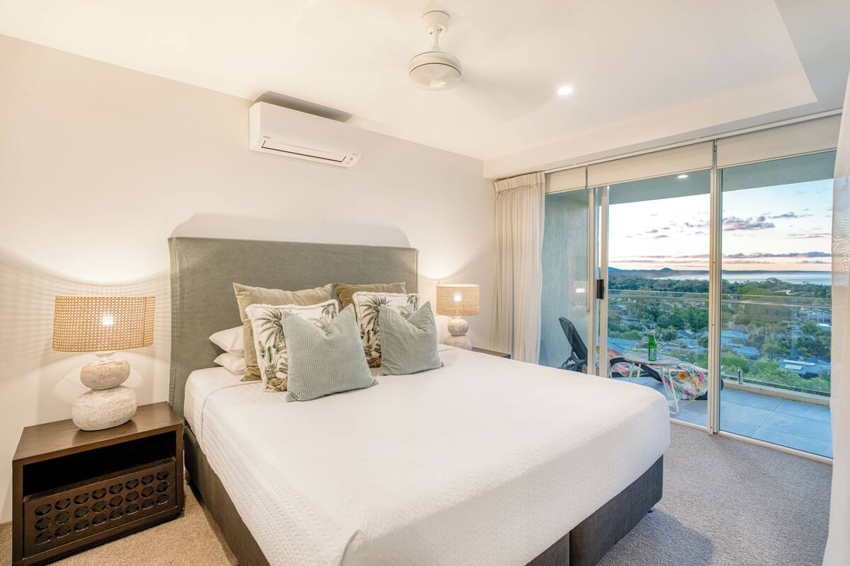 A modern bedroom with a large bed, a ceiling fan, and a balcony with a city view.