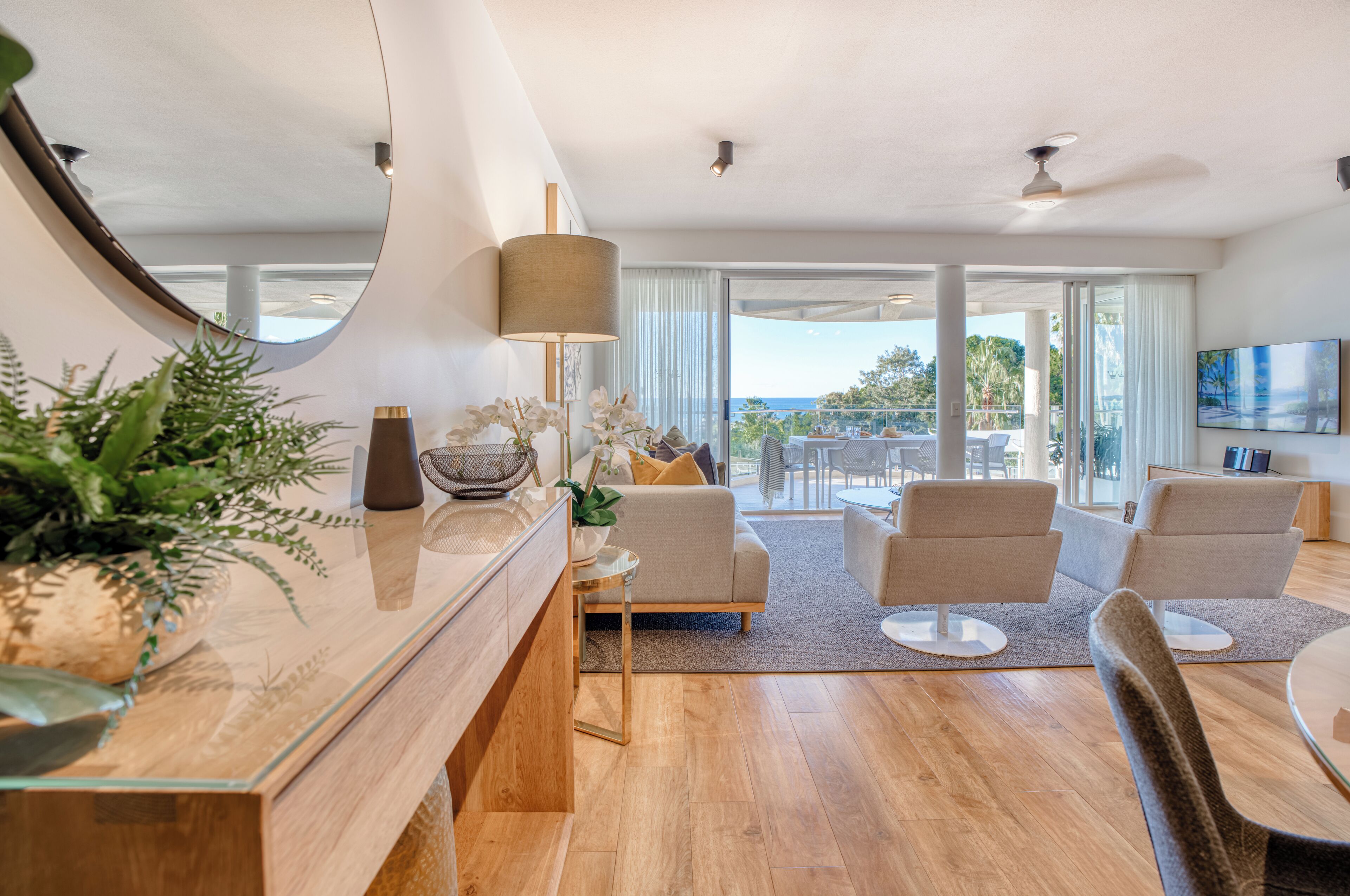 A modern living room with a glass coffee table, beige sofas, and a large window offering a view of the outdoors.