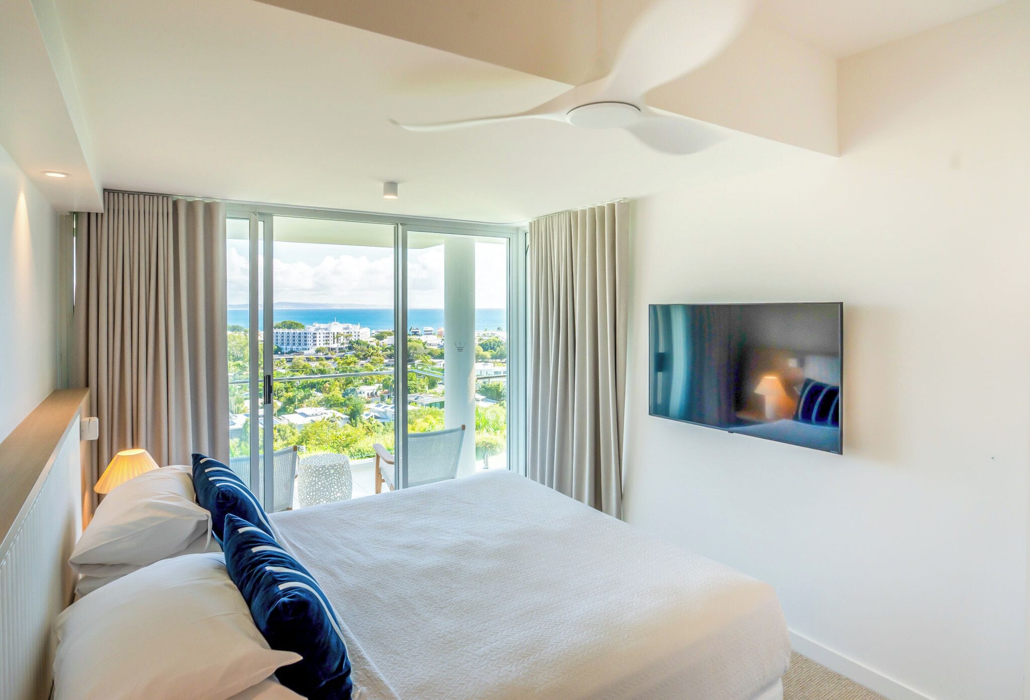 A hotel room with a bed, a TV, a ceiling fan, and a view of the city.