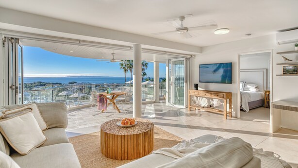 A spacious living area with a large window overlooking the ocean, a round wooden coffee table, and a television on the wall. - Noosa Crest (Noosa Heads)