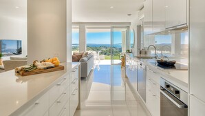 A modern kitchen with a large island, a dining area with a view, and a living room with a sofa and a TV. - Noosa Crest (Noosa Heads)