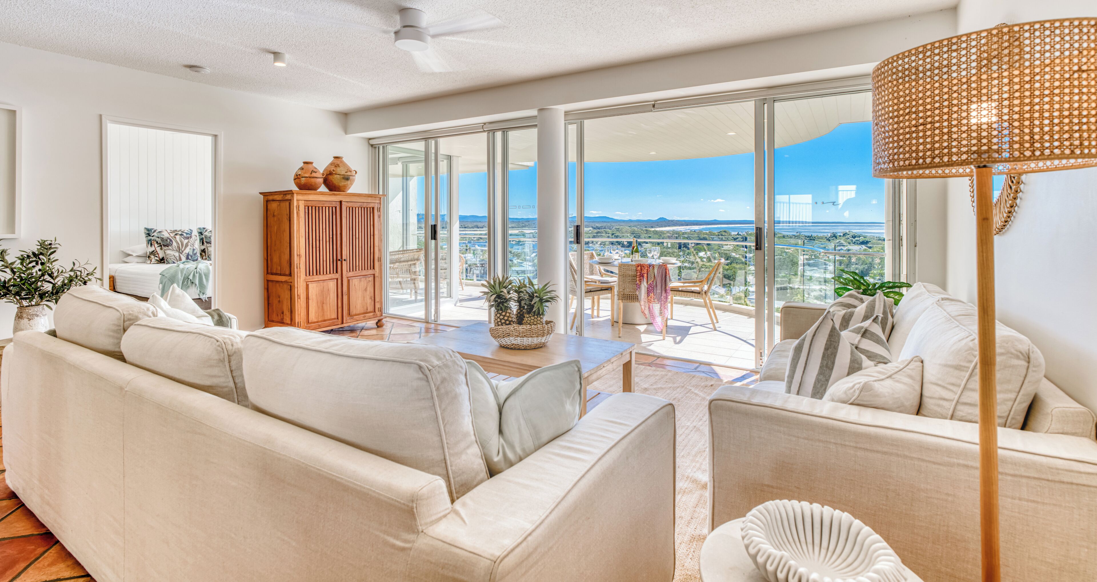 A spacious living room with a large sofa, a wooden cabinet, and a view of the city through floor-to-ceiling glass doors.