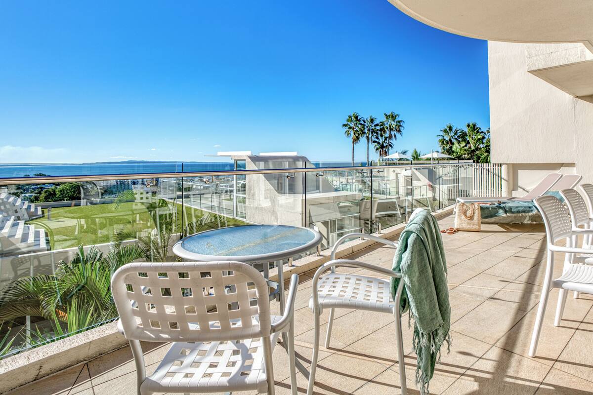 A balcony with a glass railing, white plastic chairs, a round table, and a view of the ocean and distant buildings.