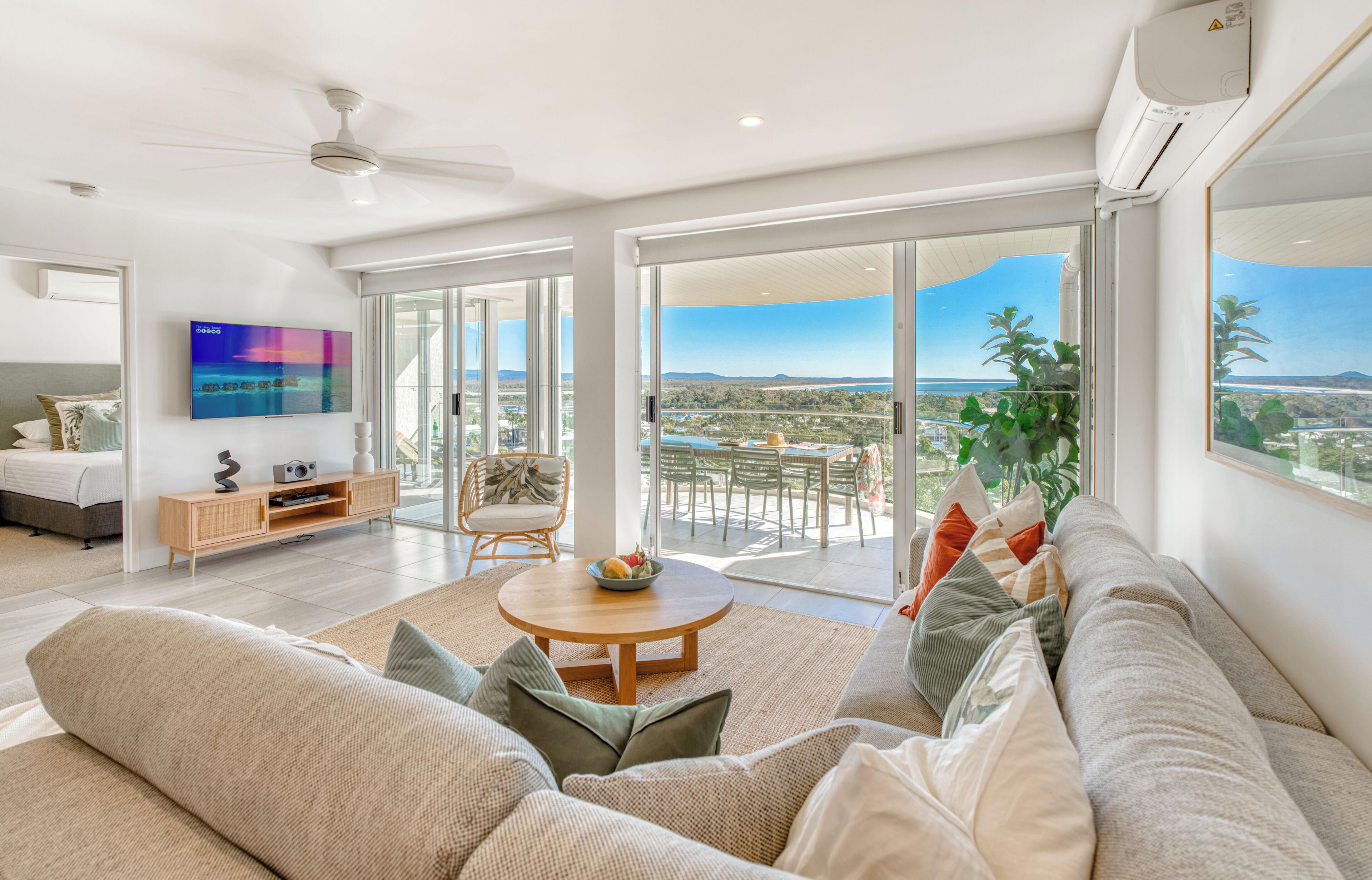 A spacious living room with a large sofa, a coffee table, and a TV mounted on the wall. There is a balcony with a view of the city and a plant on the windowsill.