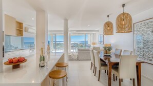 A modern kitchen with a central island, bar stools, and a dining area with a view of the city. - Noosa Crest (Noosa Heads)