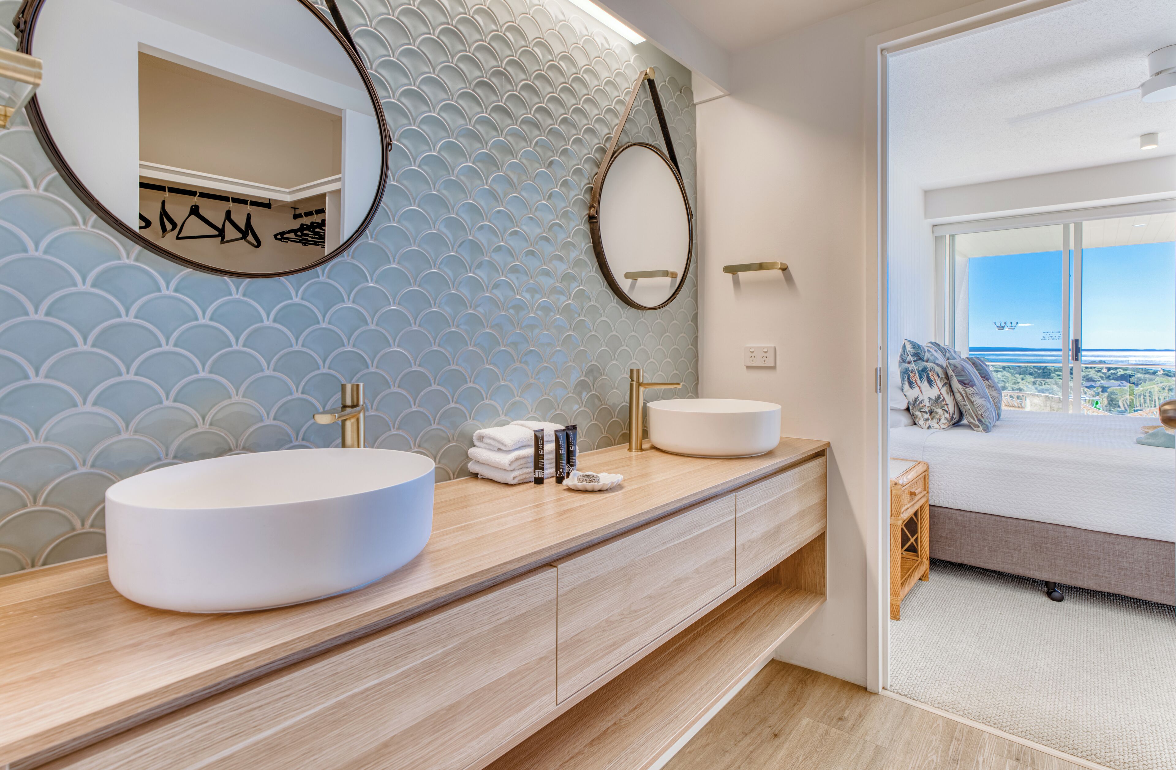 Een moderne badkamer met een groot ovaal wastafel, een spiegel, en uitzicht op een slaapkamer met een bed en een raam.