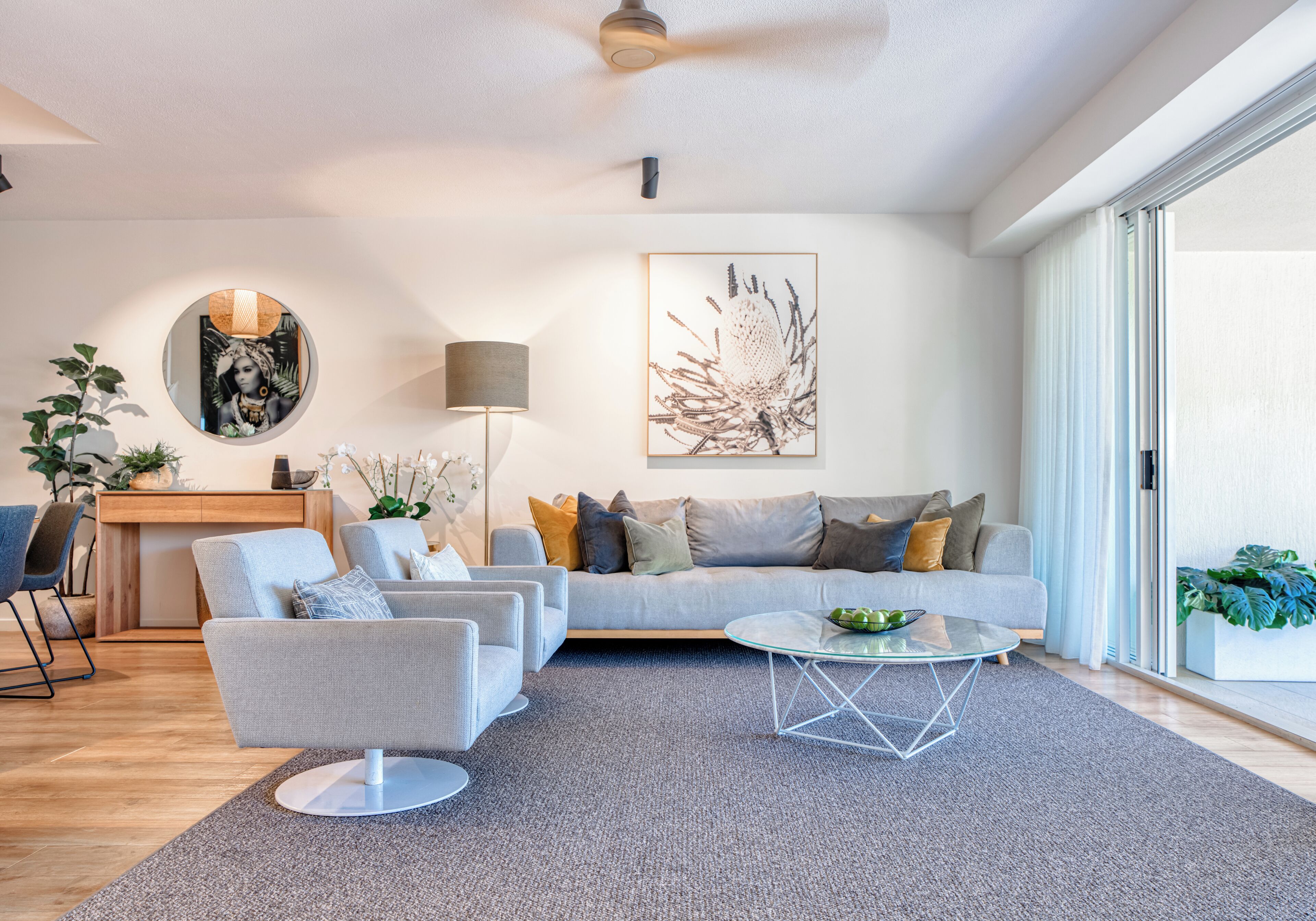 A modern living room with a grey sofa, a glass coffee table, and a grey rug. There is a desk with a lamp and a plant.