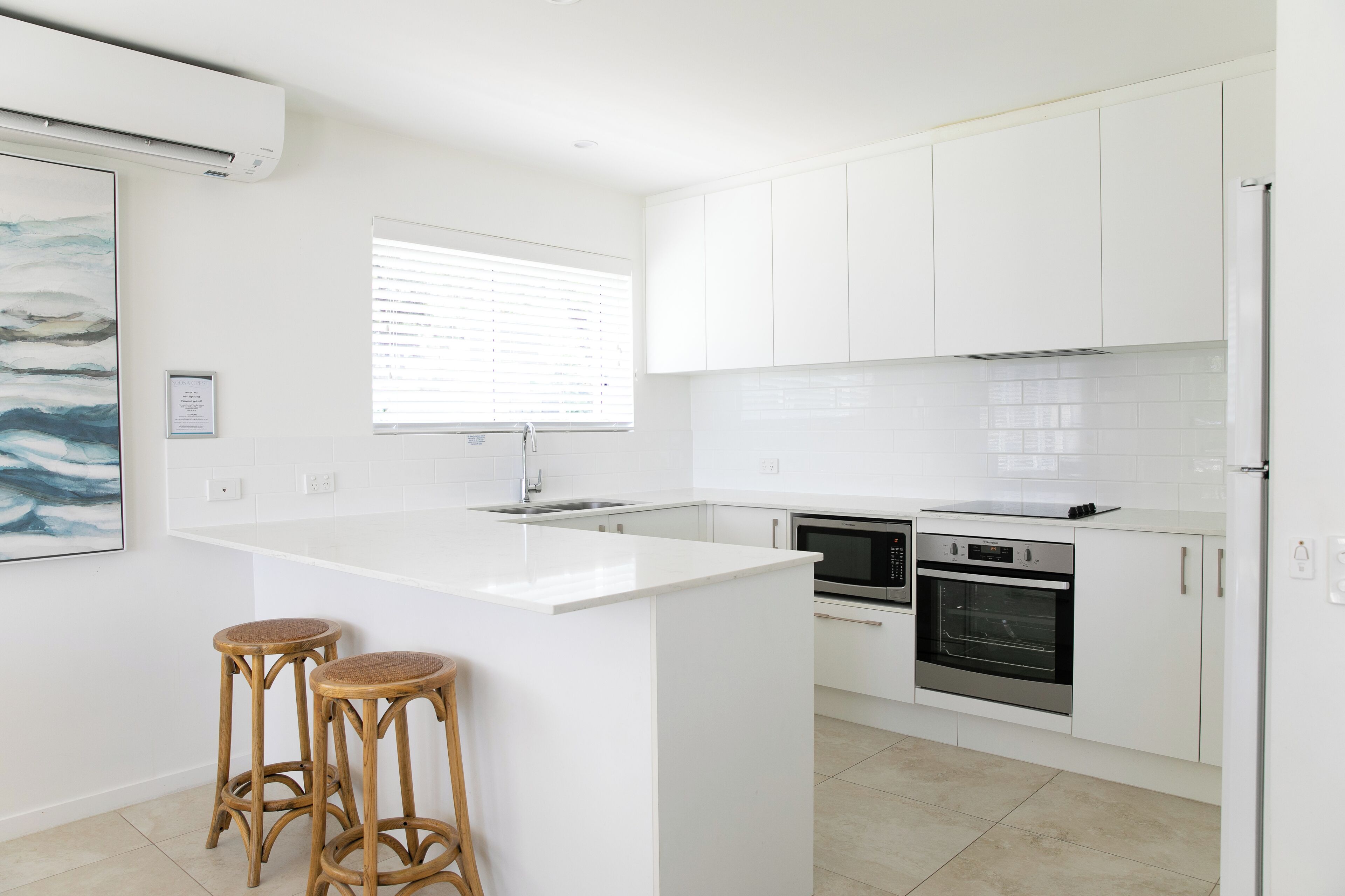 Une cuisine moderne avec des meubles blancs, un îlot central doté de tabourets, un four encastré et une fenêtre avec des stores.
