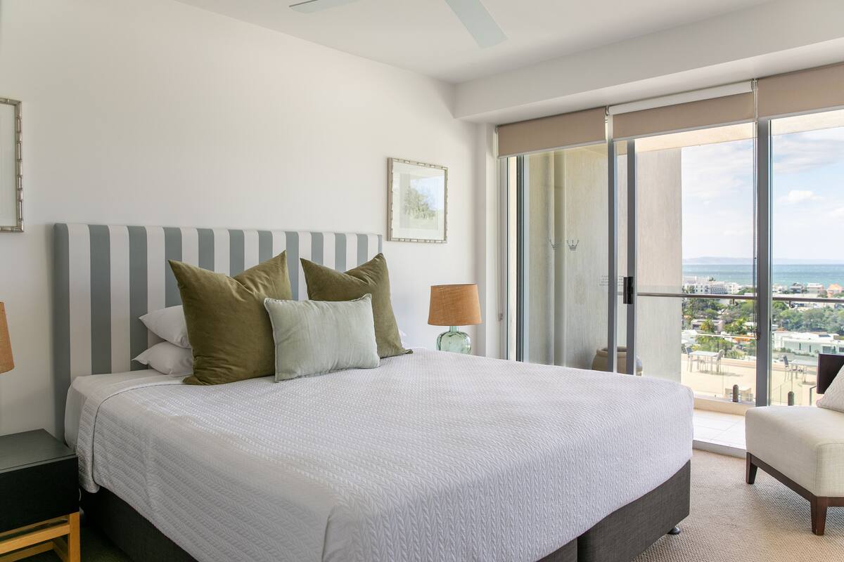 A spacious bedroom with a large bed, a view of the city through floor-to-ceiling windows, and a balcony with a chair.