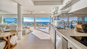 A modern kitchen with a view of the ocean, featuring a dining area, large windows, and a high ceiling. - Noosa Crest (Noosa Heads)