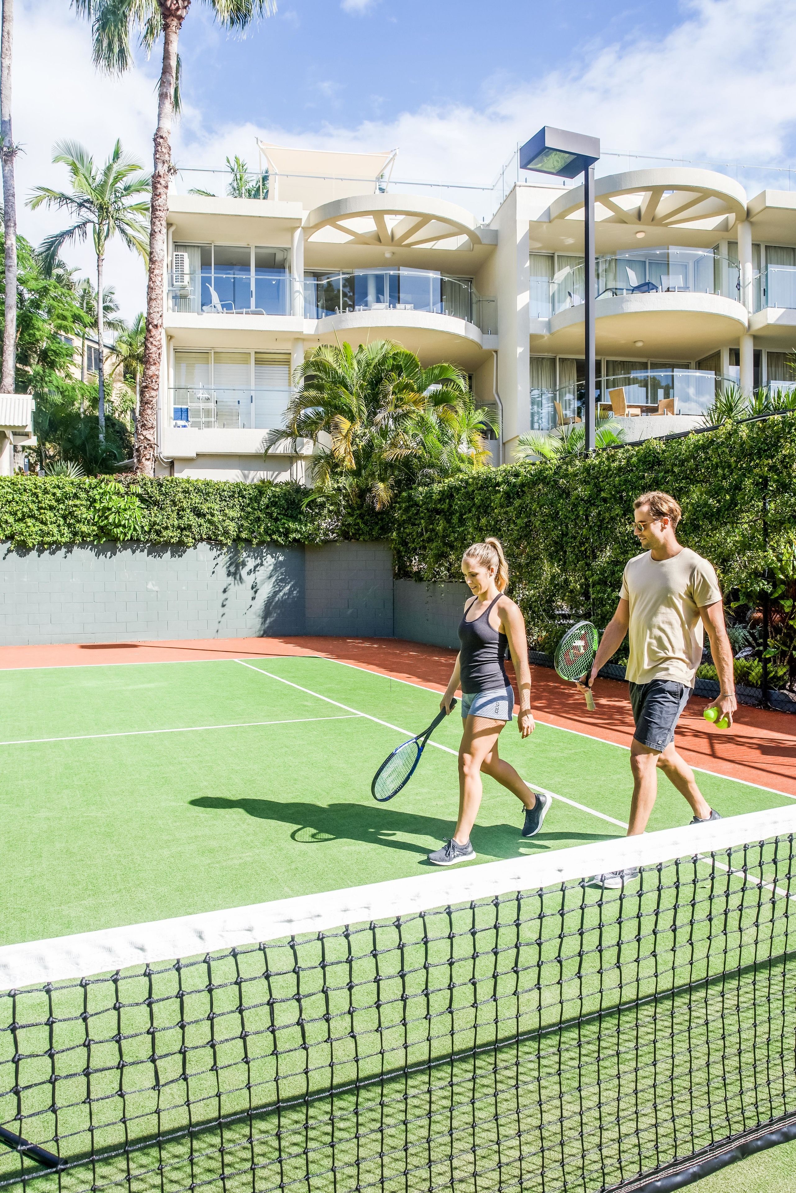 A tennis court with a net, surrounded by a building with balconies and a hedge.