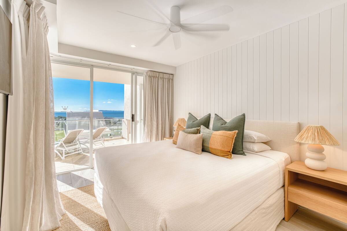 A bedroom with a large bed, a ceiling fan, a nightstand, and a view of the ocean through a sliding glass door.