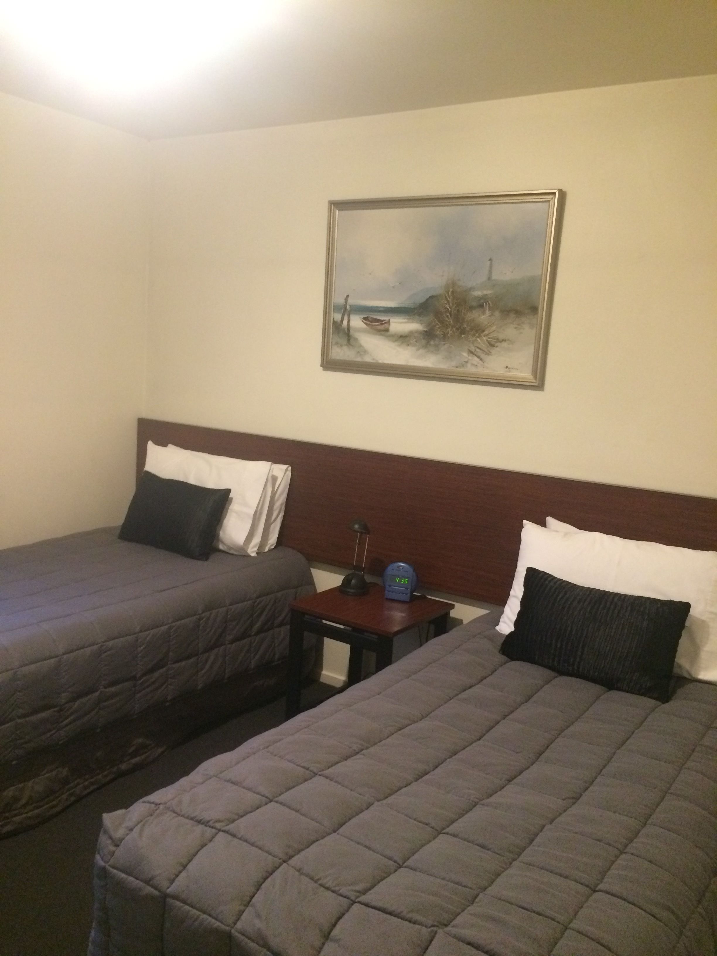 A hotel room with two beds, a nightstand with a lamp and alarm clock, and a painting on the wall.