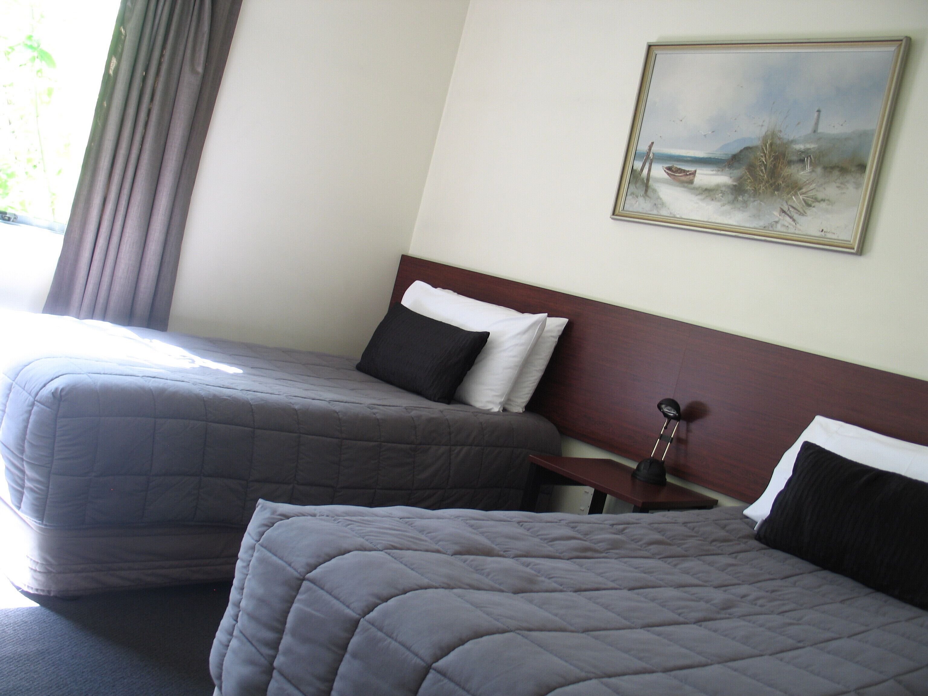 A hotel room with two beds, a painting of a beach scene, and a lamp on a bedside table.