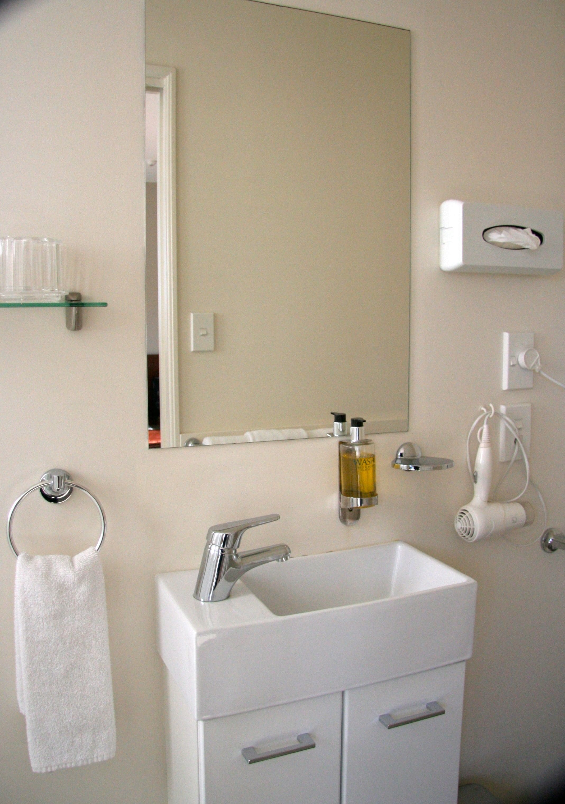 A bathroom with a white sink, a mirror, a towel ring, a glass shelf, and a hairdryer.