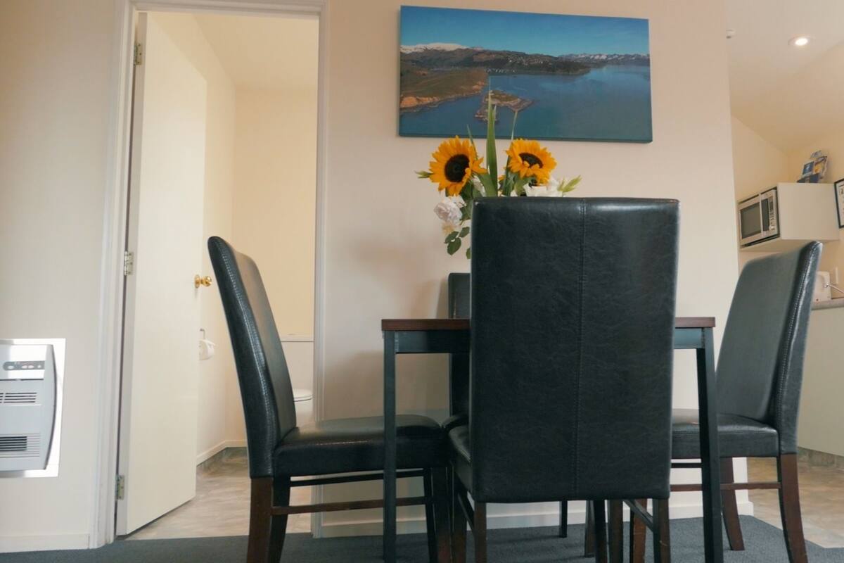A dining area with a table and four chairs, a painting of a landscape on the wall, and a kitchen area with a microwave.