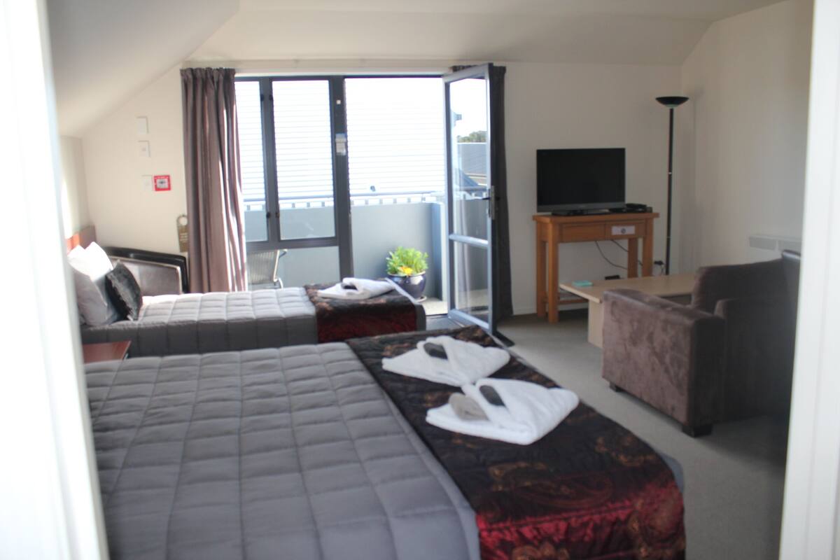 A hotel room with two beds, a sofa, a TV, and a balcony with a plant.