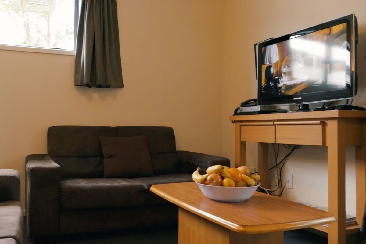 A living room with a brown leather sofa, a television on a wooden stand, and a bowl of fruit on a small table.