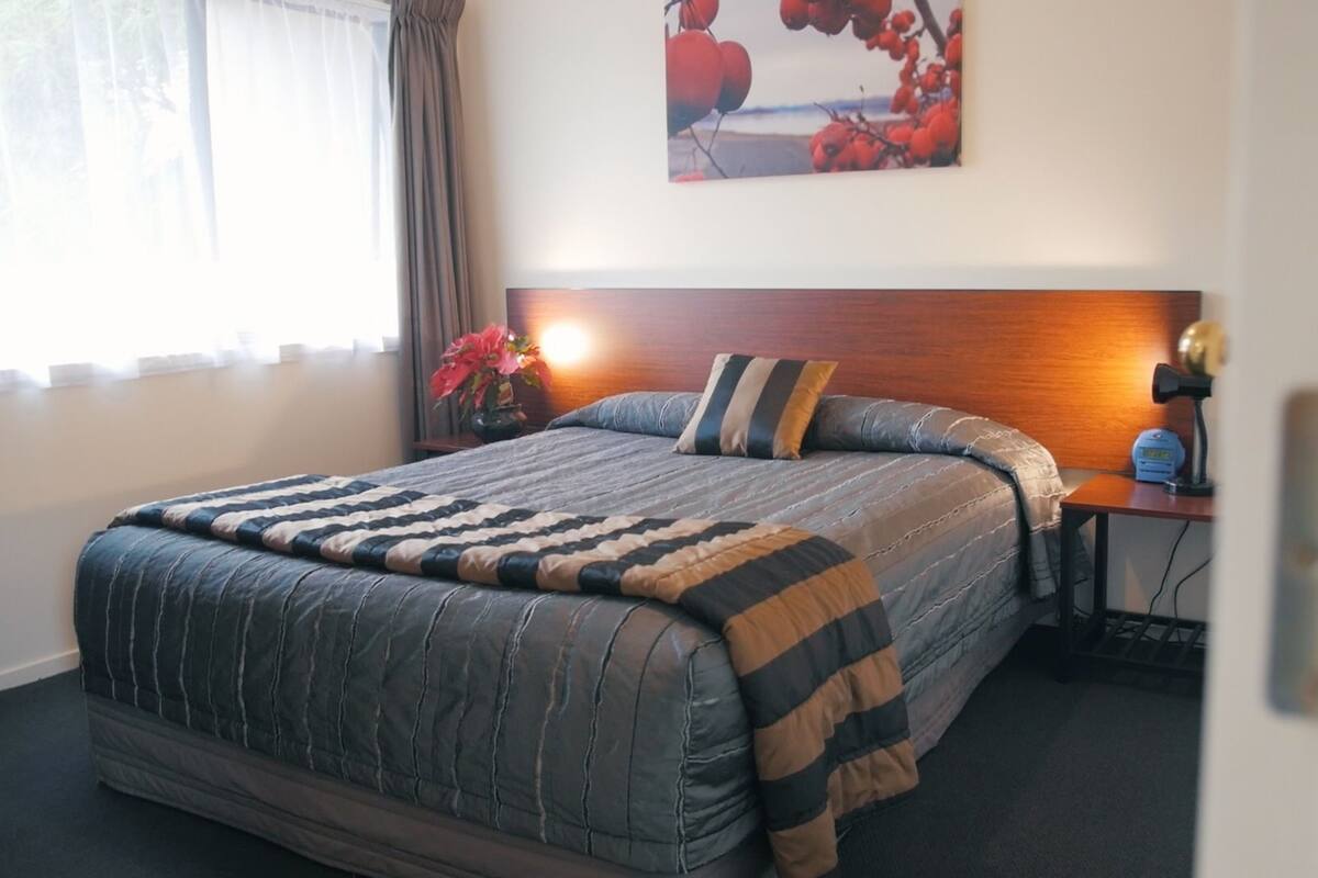 A neatly made bed with a striped blanket, a bedside table with a lamp, and a painting of red berries on the wall.