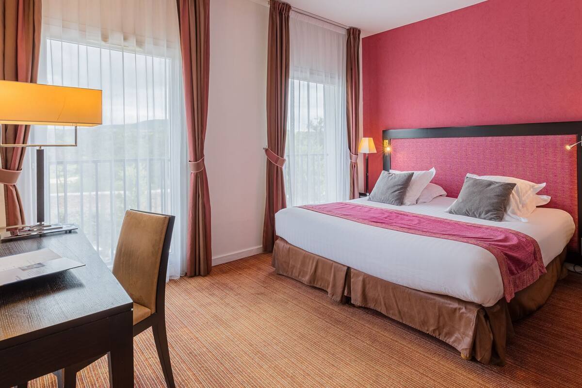 Habitación de hotel con cama, lámparas de noche, escritorio y silla, y ventana con cortinas.