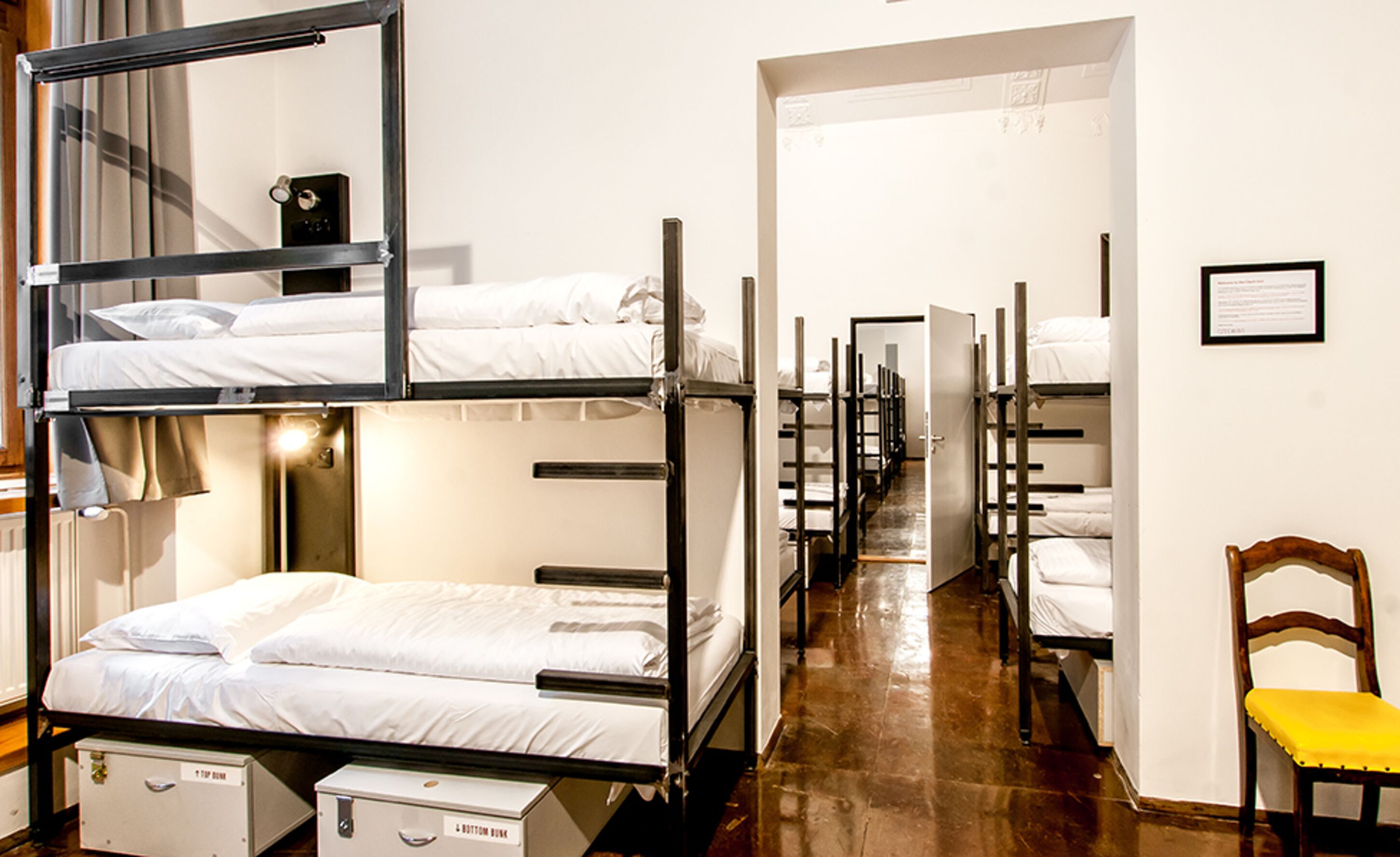 A dormitory room with bunk beds, a wooden chair, and a framed certificate on the wall.