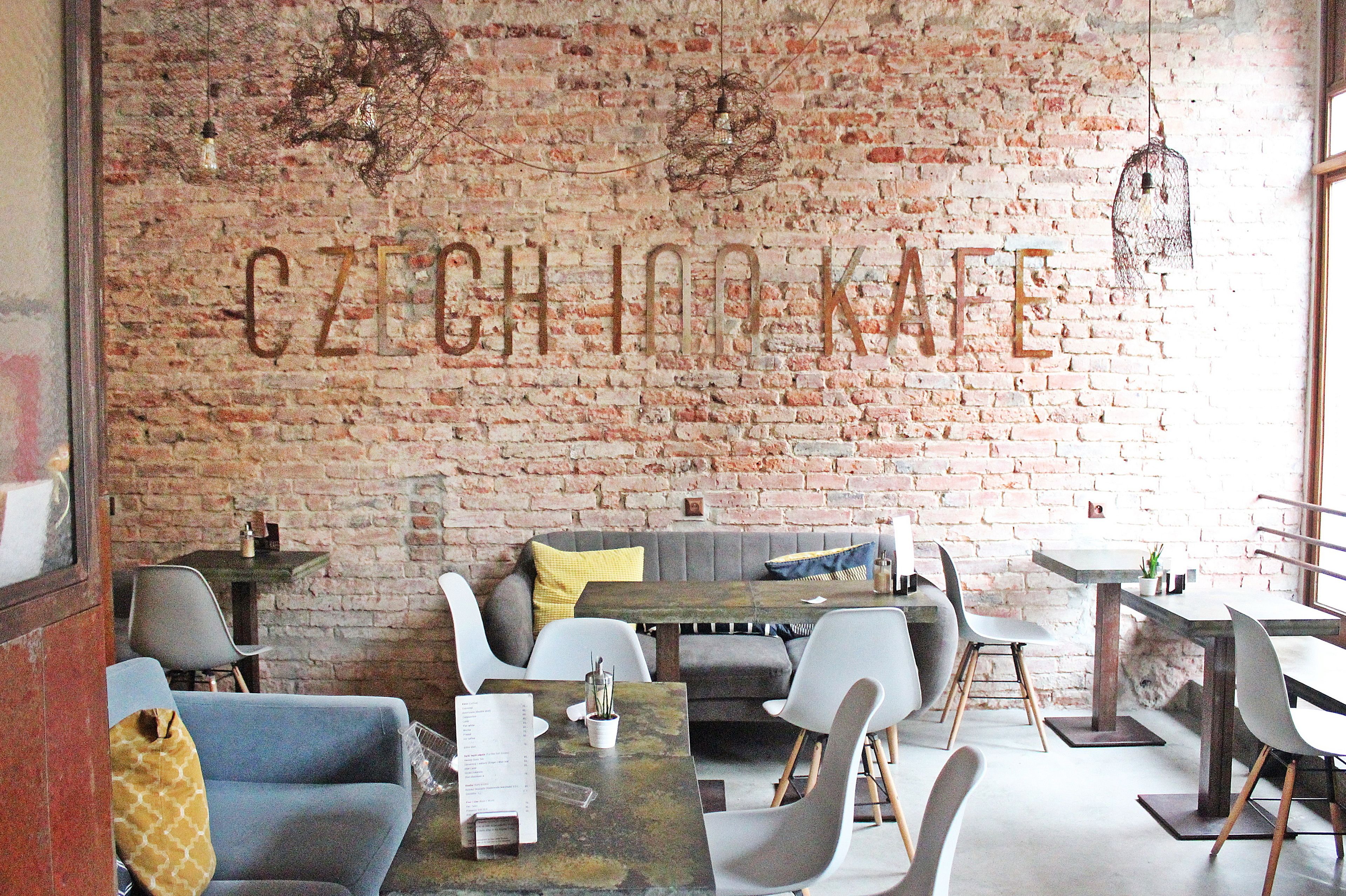 A cafe with a brick wall and the words "CZECH INKAKE" in large letters. There are tables and chairs arranged for seating.