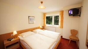 A hotel room with a double bed, bedside tables with lamps, a TV mounted on the wall, a window with curtains, and a small chair. - Appartements Furstauer (Saalbach-Hinterglemm)