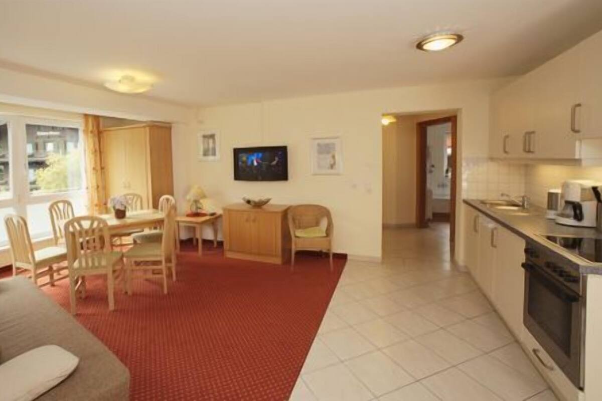 A living room with a red carpet, a dining area with wooden furniture, a kitchen with a stove and coffee maker, and a TV on the wall.
