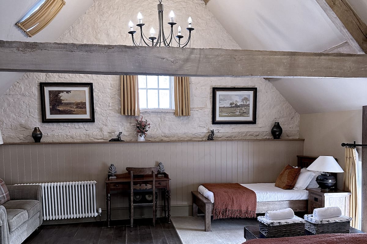 A cozy attic bedroom with a sloped ceiling, wooden beams, a chandelier, two beds, a sofa, a table, and a radiator.