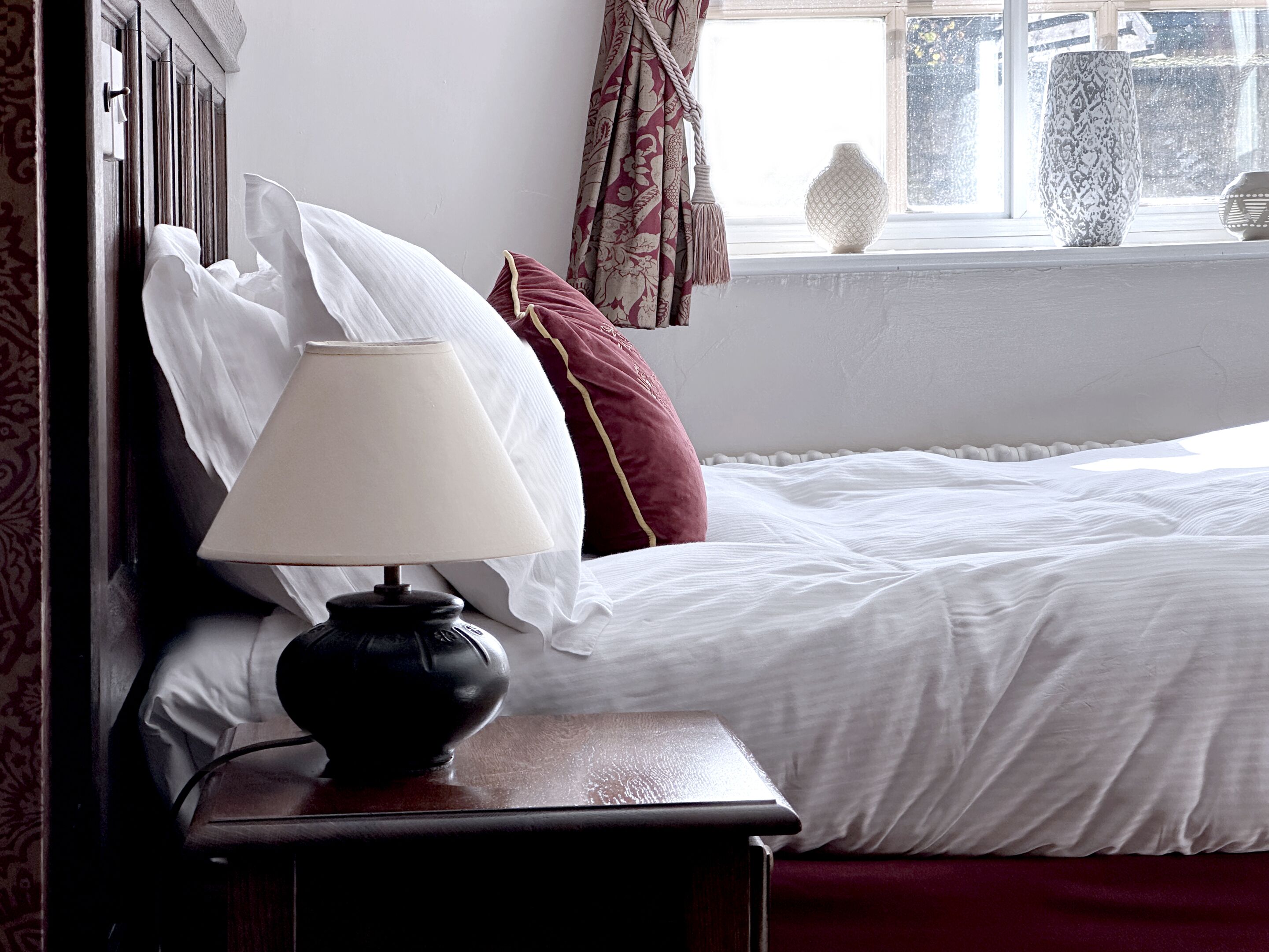 A neatly made bed with white linens, a dark wooden nightstand with a lamp, and a window with curtains.