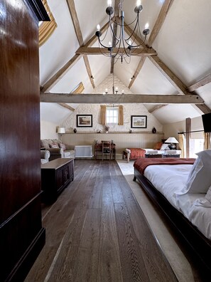 A spacious attic bedroom with exposed wooden beams, a chandelier, a large bed, a desk, and a radiator. - The Crown at Marnhull (Sturminster Newton)
