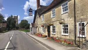 A traditional stone building with a sign reading "The Crown." It features a road with a speed limit sign, flower beds, and a church in the background. - The Crown at Marnhull (Sturminster Newton)