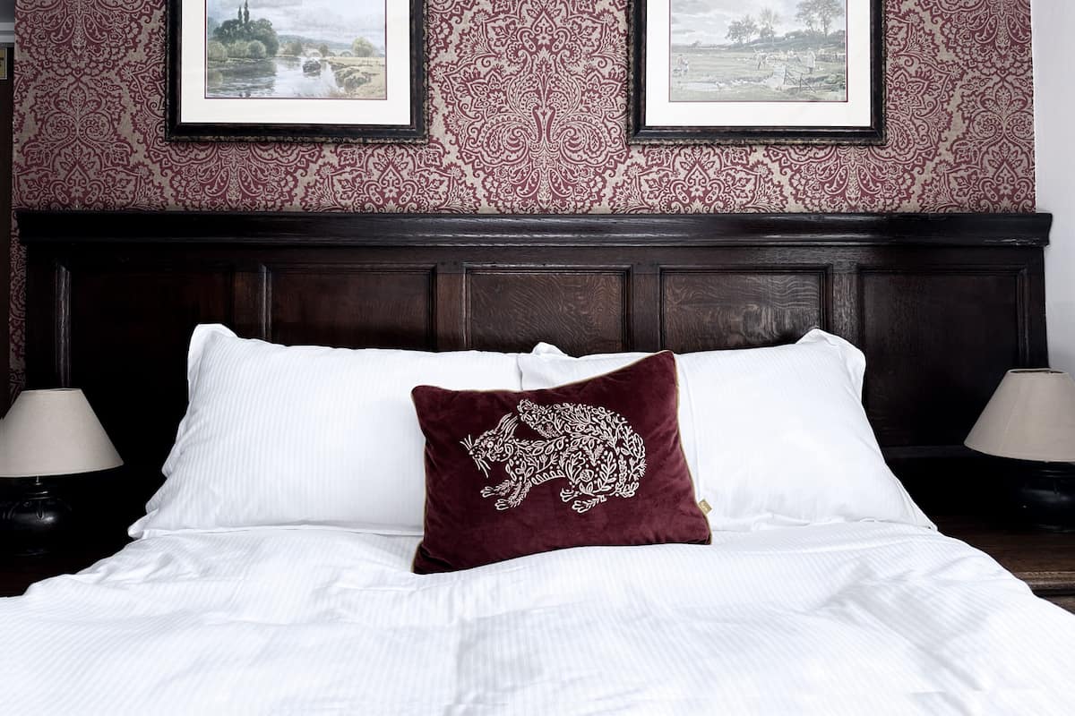 A neatly made bed with white linens and a single decorative pillow, flanked by two bedside lamps. Above the bed, there are two framed pictures on a patterned wallpaper.