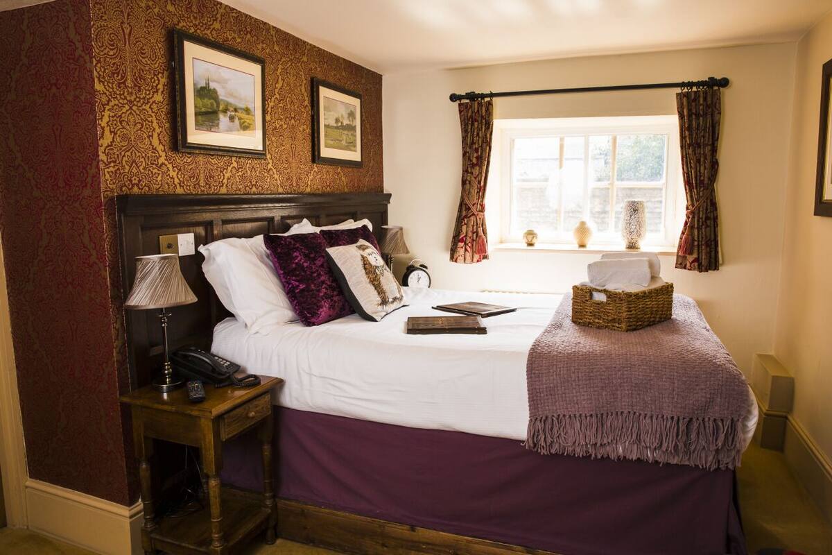 A neatly arranged bedroom with a large bed, a bedside table with a lamp, a window with curtains, and two framed pictures on the wall.
