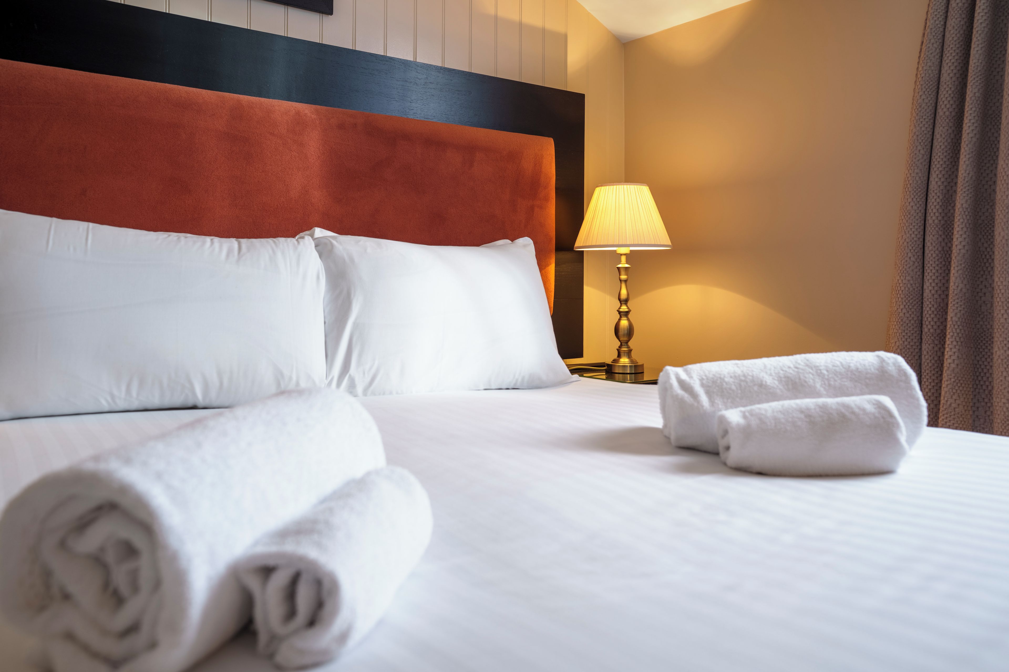 A hotel room with a neatly made bed, a bedside lamp, and a rolled towel on the bed.