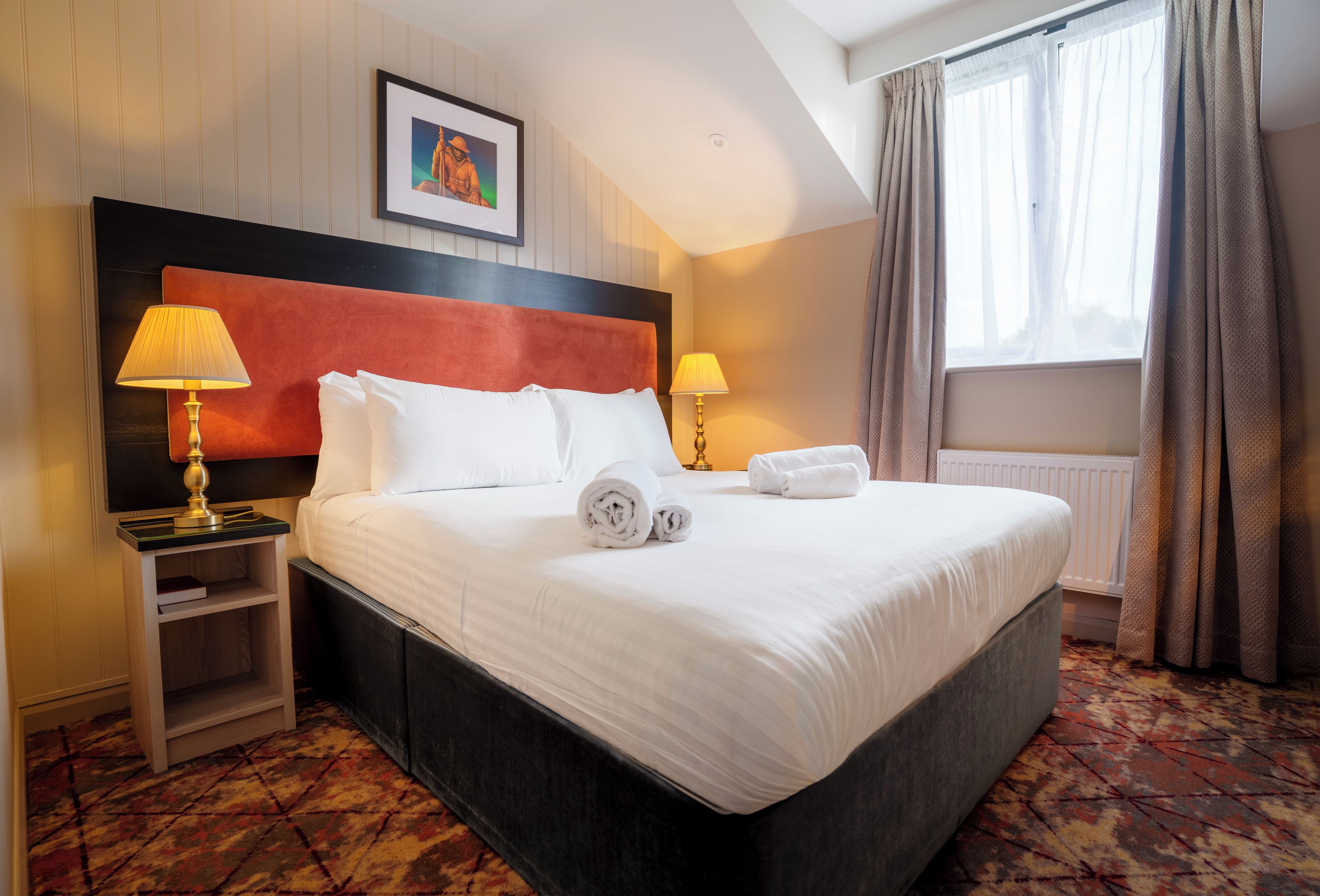 Habitación de hotel con una cama grande, dos lámparas de noche, un cuadro en la pared y una ventana con cortinas.