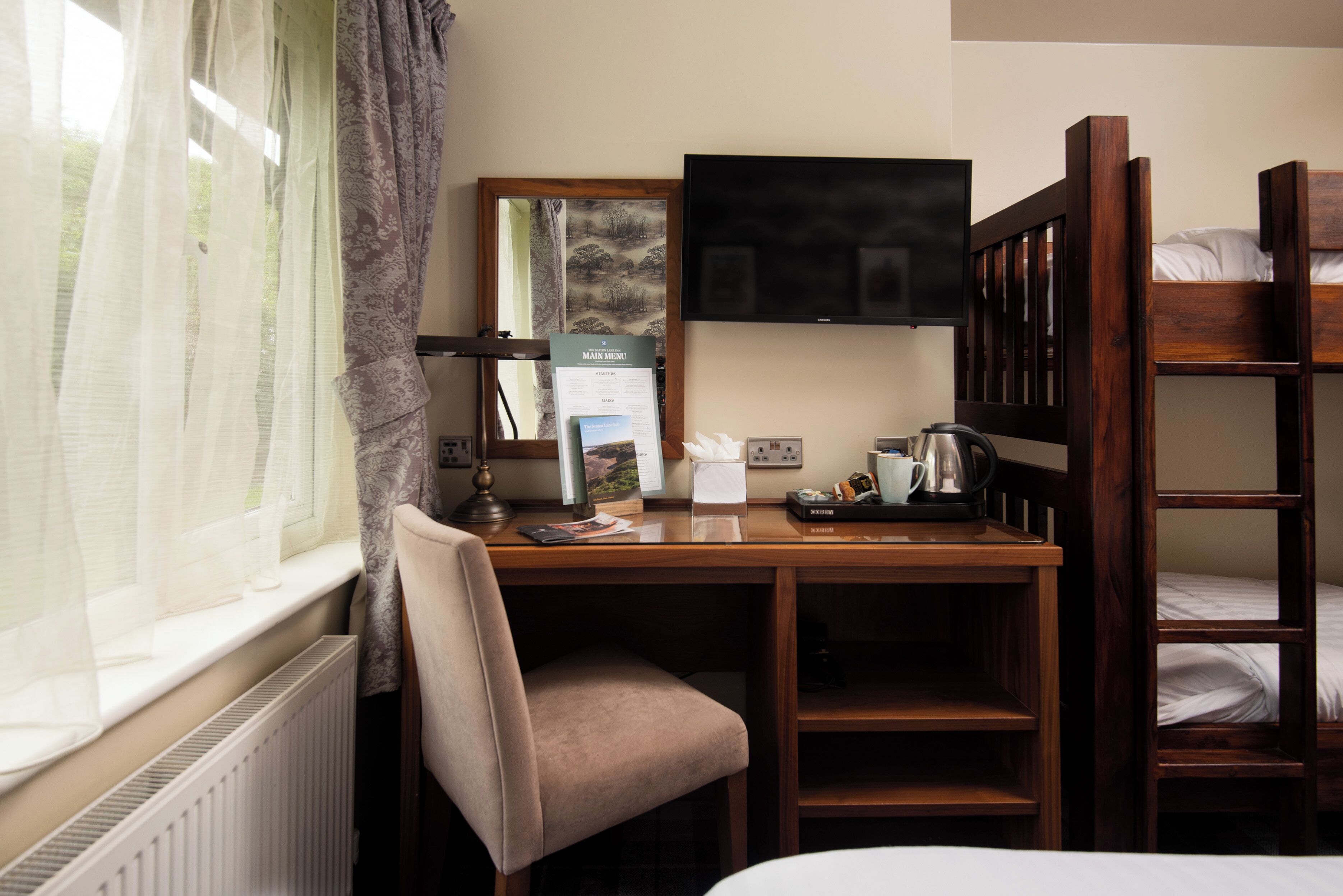 Une chambre d’hôtel avec un bureau en bois, une chaise, un lit superposé, une télévision et une fenêtre avec des rideaux légers.