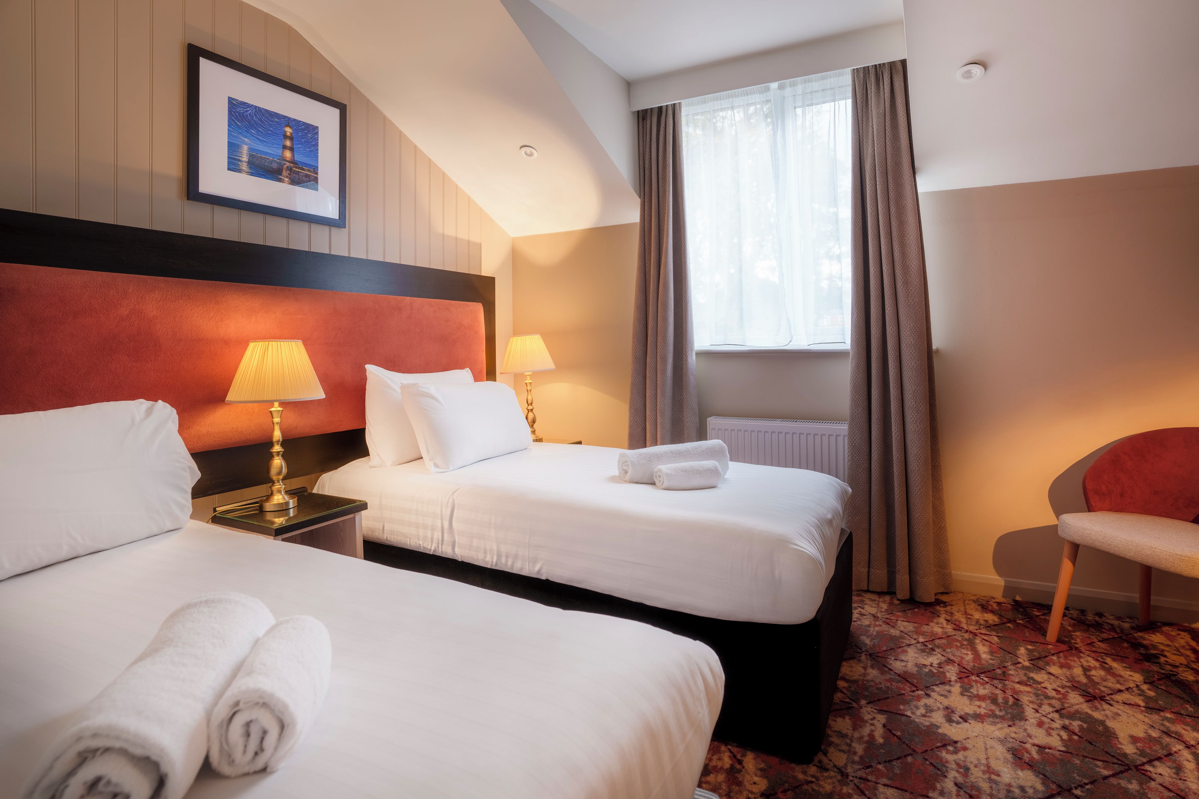 Habitación de hotel con dos camas, cabecera roja, dos lámparas de noche, ventana con cortinas, radiador y una silla.