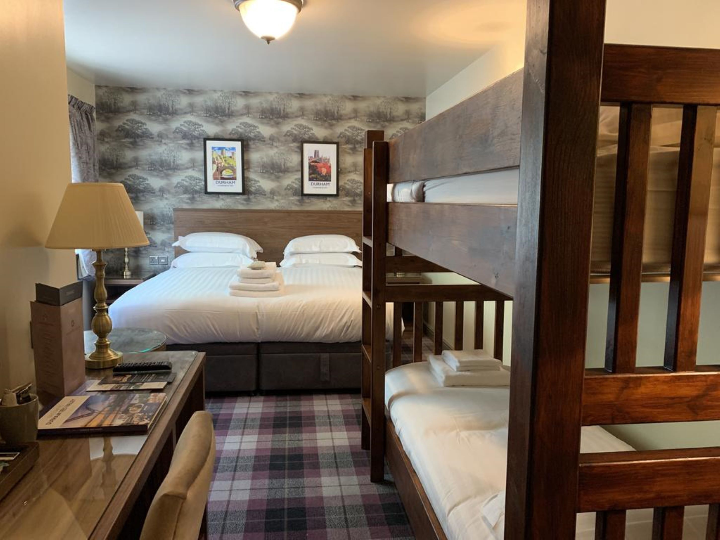 A hotel room with a bunk bed, a desk with a lamp, a checkered floor, and framed pictures on the wall.