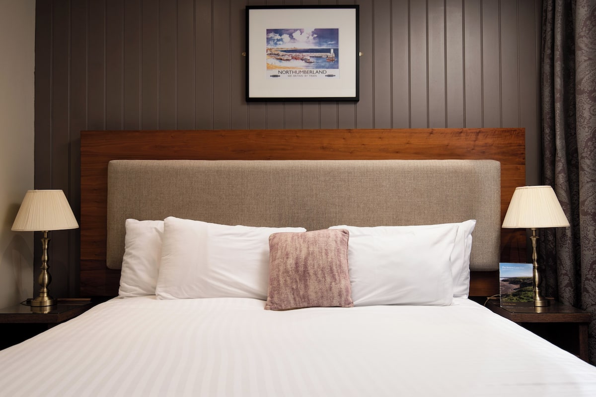 A neatly made bed with white linens and a single pink throw pillow, flanked by two bedside lamps on wooden nightstands. A framed picture of Northumberland hangs above the bed.