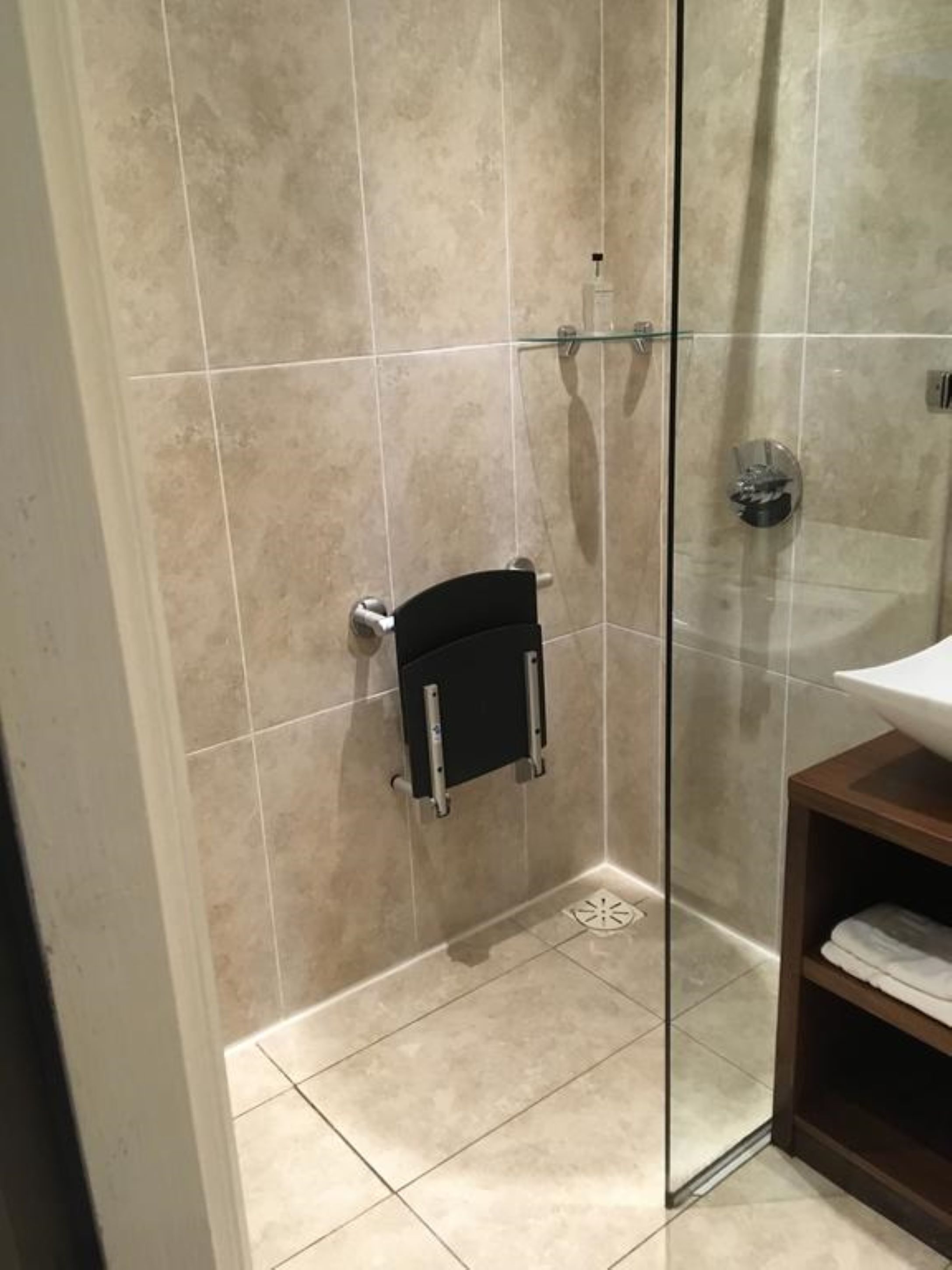 Un baño moderno con cabina de ducha de vidrio, azulejos color beige y un mueble de tocador de madera con lavamanos blanco.