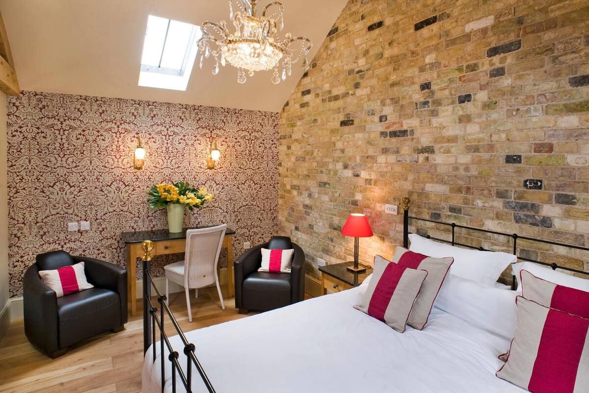 A bedroom with a large bed, two chairs, a desk, a chandelier, and a brick wall.