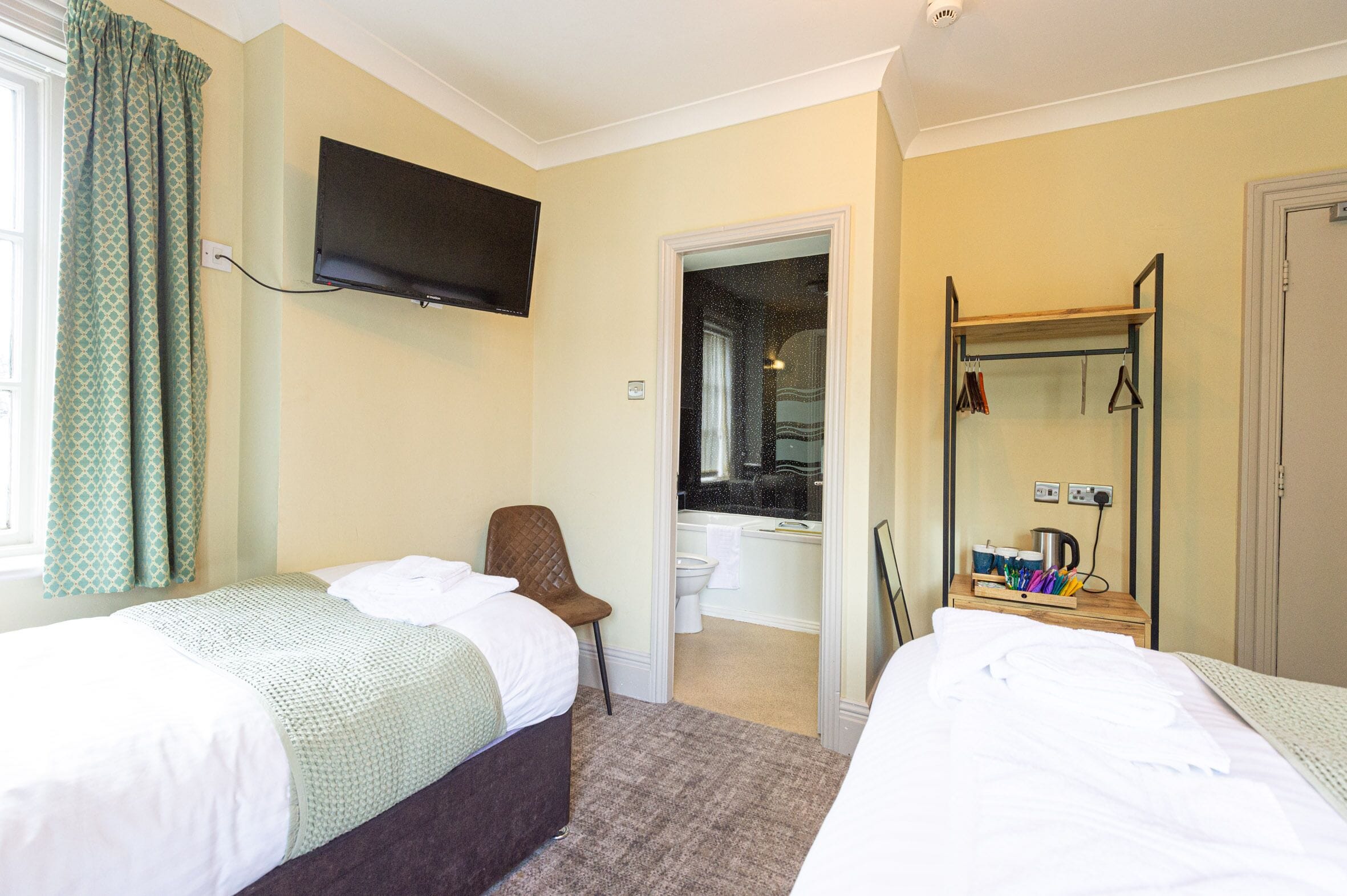 Habitación de hotel con dos camas, un televisor montado en la pared, una silla, una mesita con tetera y tazas, y un espejo con ropa colgada.